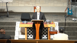 Centered shot of a pastor speaking at a wooden pulpit in a well-lit church sanctuary, full stage visible.