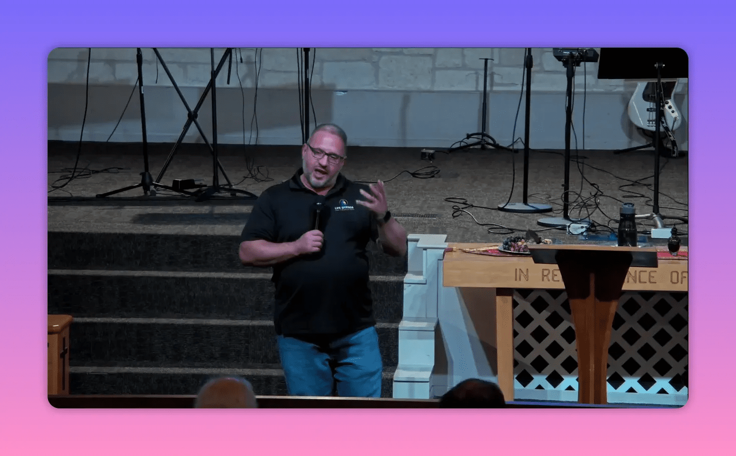 Speaker on stage making an expressive hand gesture while holding a microphone, communion table visible to the right