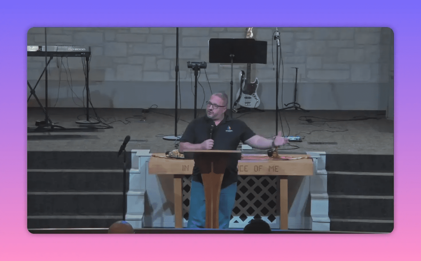 Church speaker at a lectern with an outstretched arm, stage instruments and communion table visible — a wide, clear sermon frame