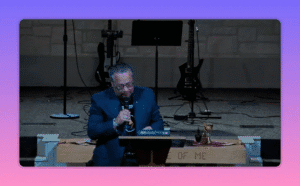 Church leader leaning over a pulpit with an open Bible visible and communion cup to the side, stage guitars behind.
