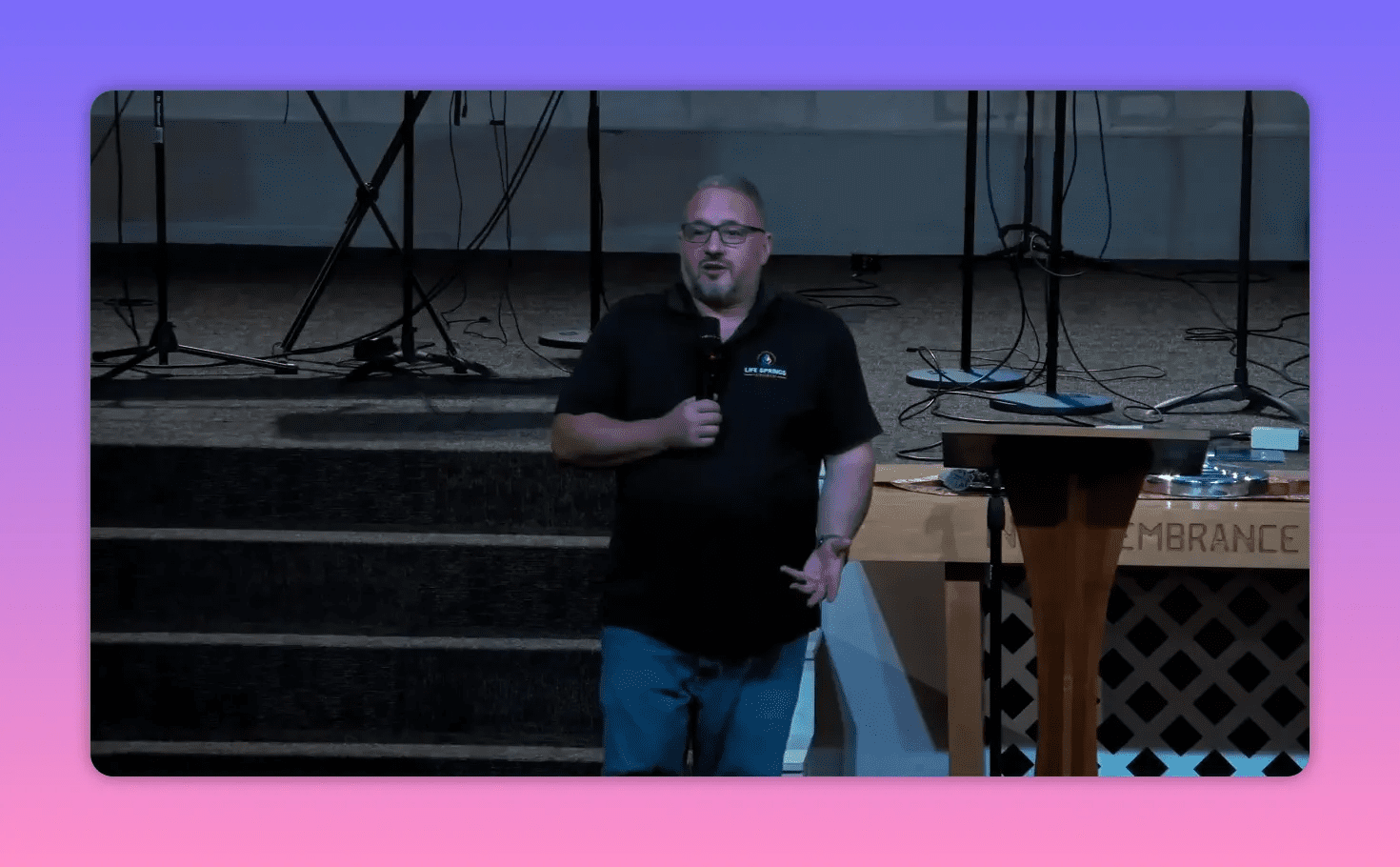 Centered mid-shot of a speaker holding a microphone beside the communion table, addressing the congregation with clear lighting and focus.