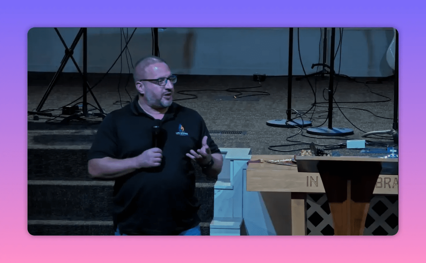 Church speaker gesturing with one hand while holding a microphone beside the pulpit; stage setup and microphones visible