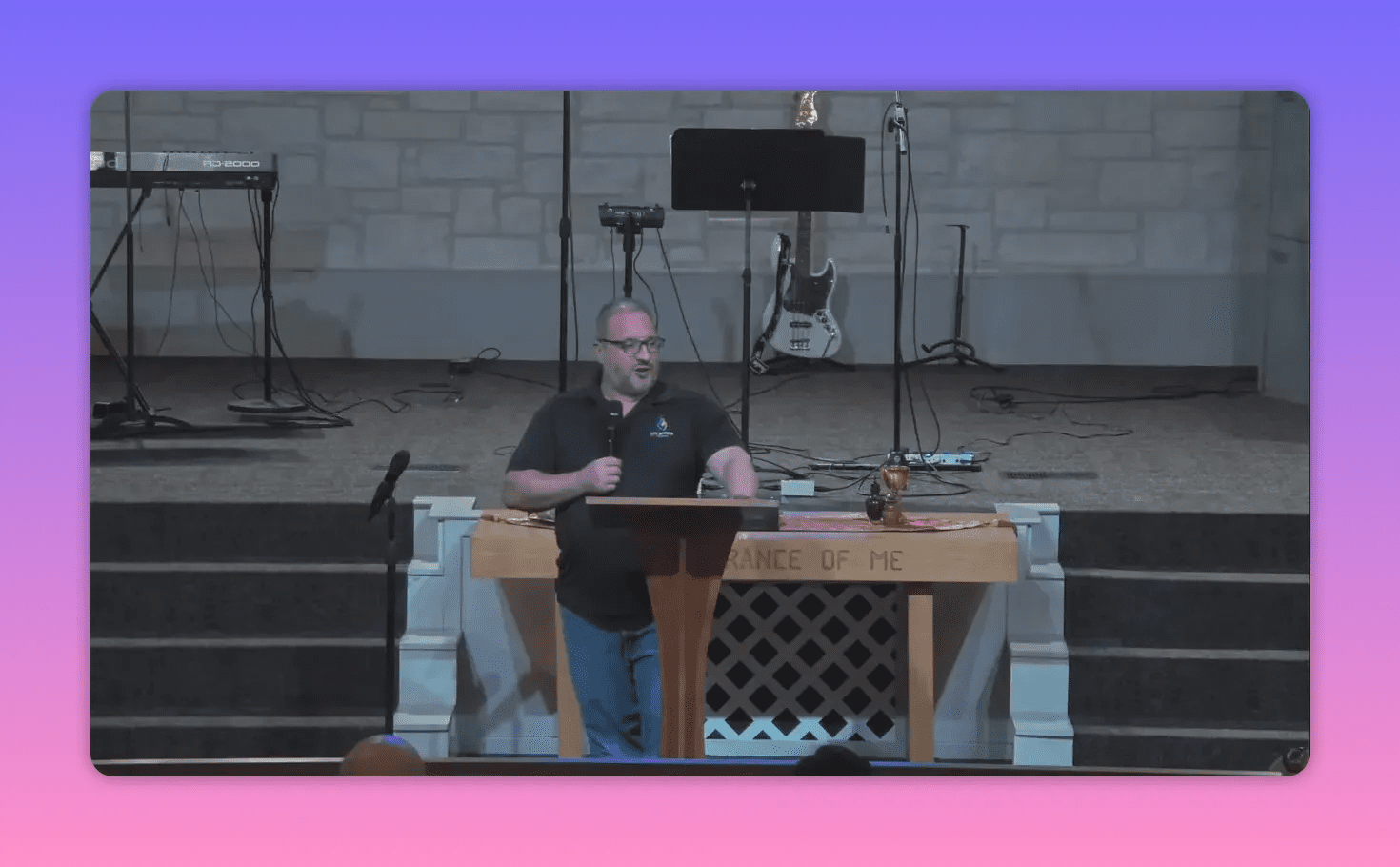 Church speaker at a lectern with one hand resting on the pulpit and the other holding a microphone, communion items and instruments visible behind