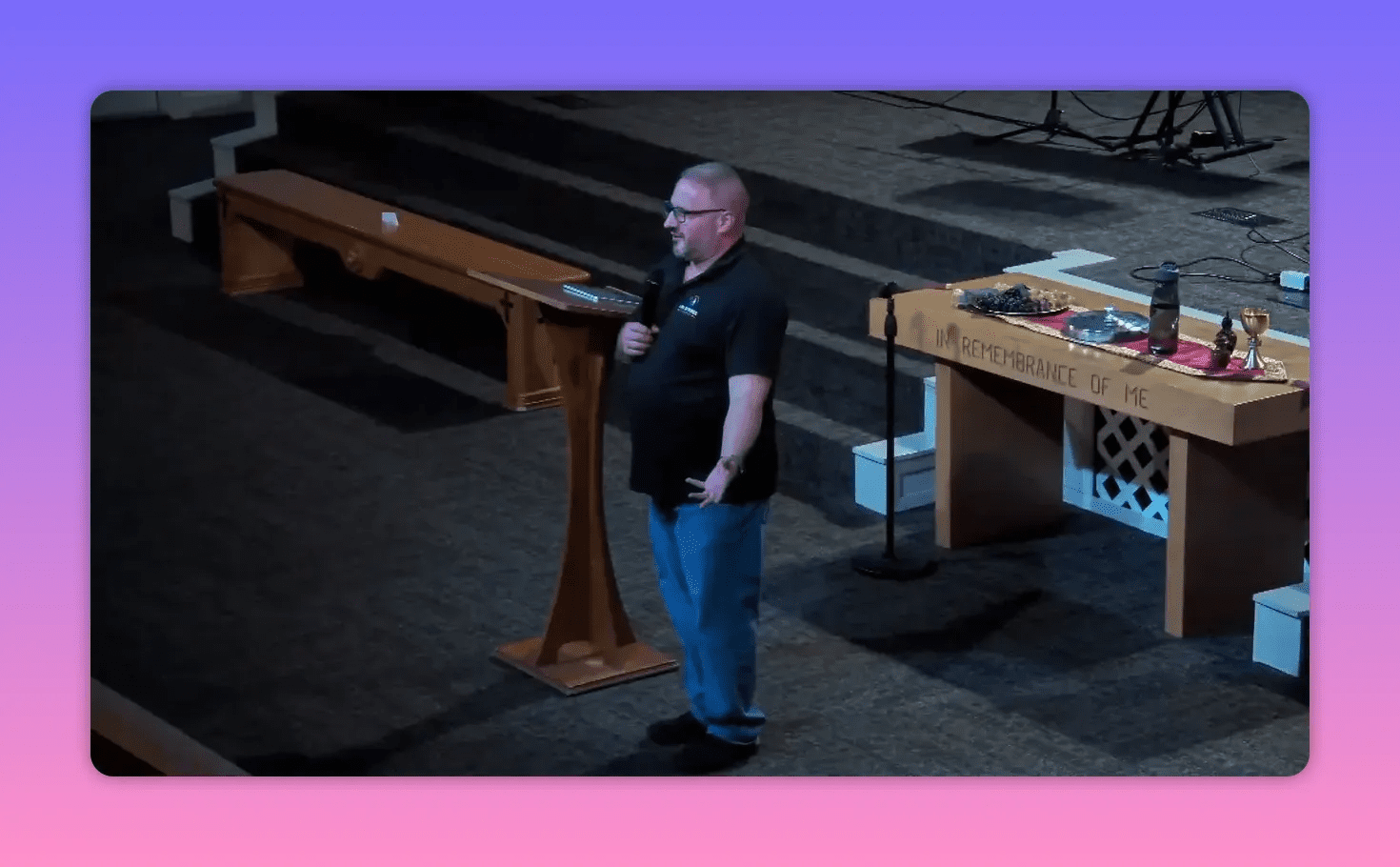 Clear shot of the pastor with an open-hand gesture standing beside the communion table engraved 'In Remembrance of Me' on stage.