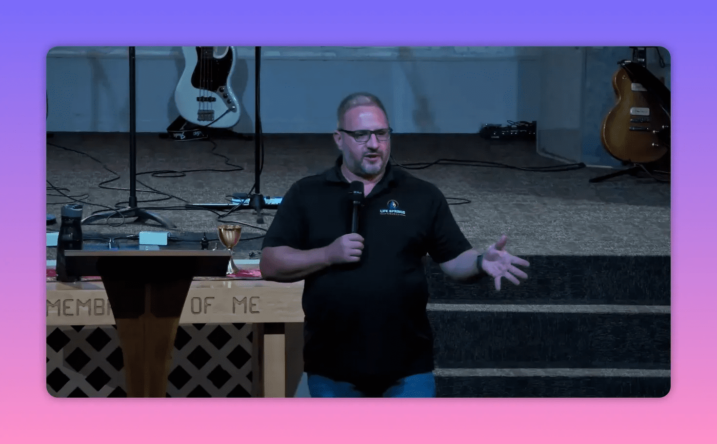 Clear frontal shot of a speaker holding a microphone with an open-hand gesture, lectern and communion table visible