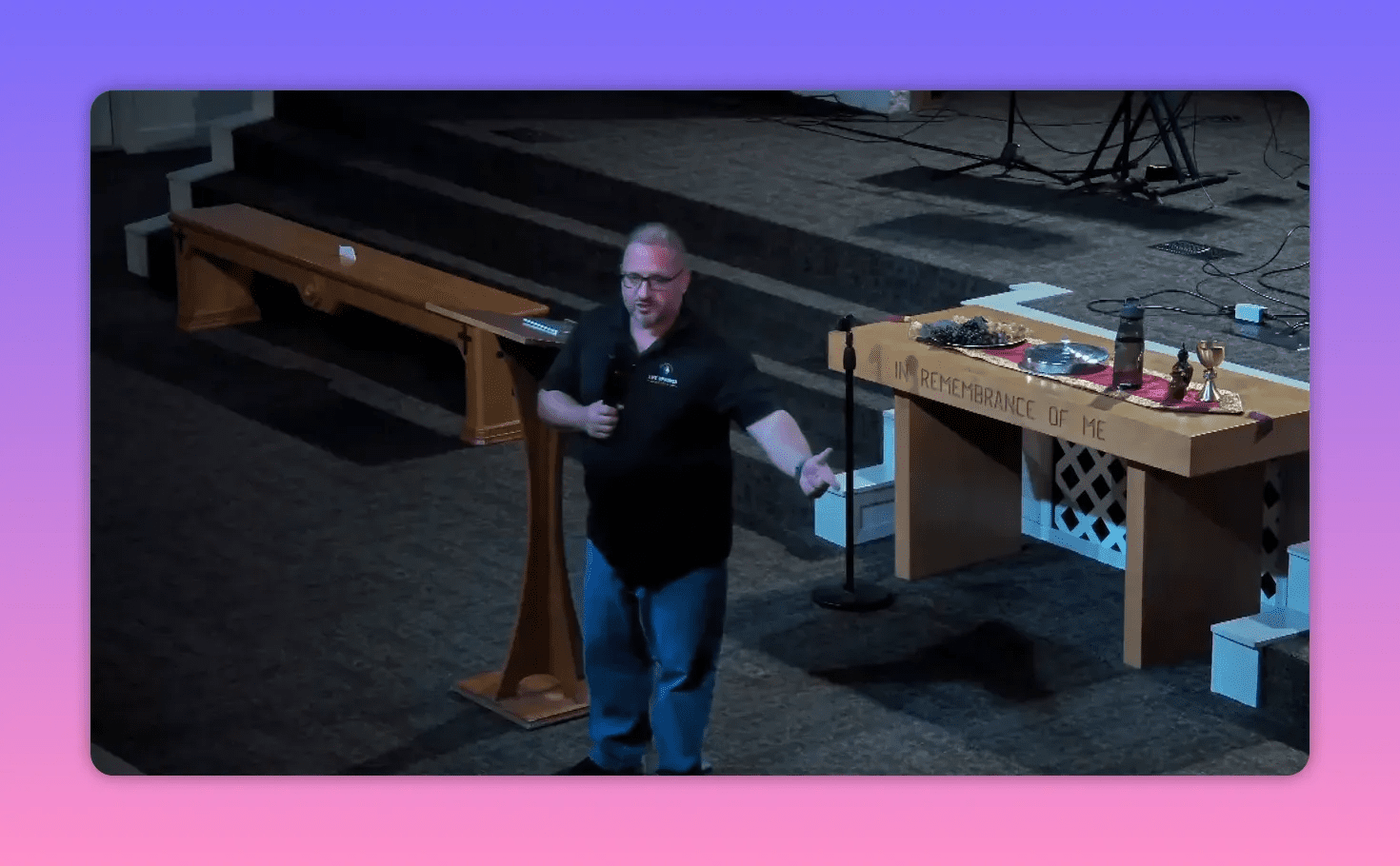 Clear wide shot of the speaker with an open hand gesture beside the communion table engraved 'In Remembrance of Me', showing stage and steps behind.