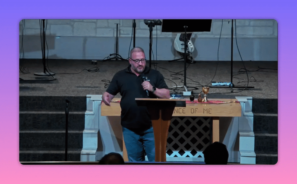 Speaker centered at the pulpit gesturing while holding a microphone, communion table and chalice visible behind him
