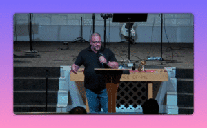 Speaker centered at the pulpit gesturing while holding a microphone, communion table and chalice visible behind him
