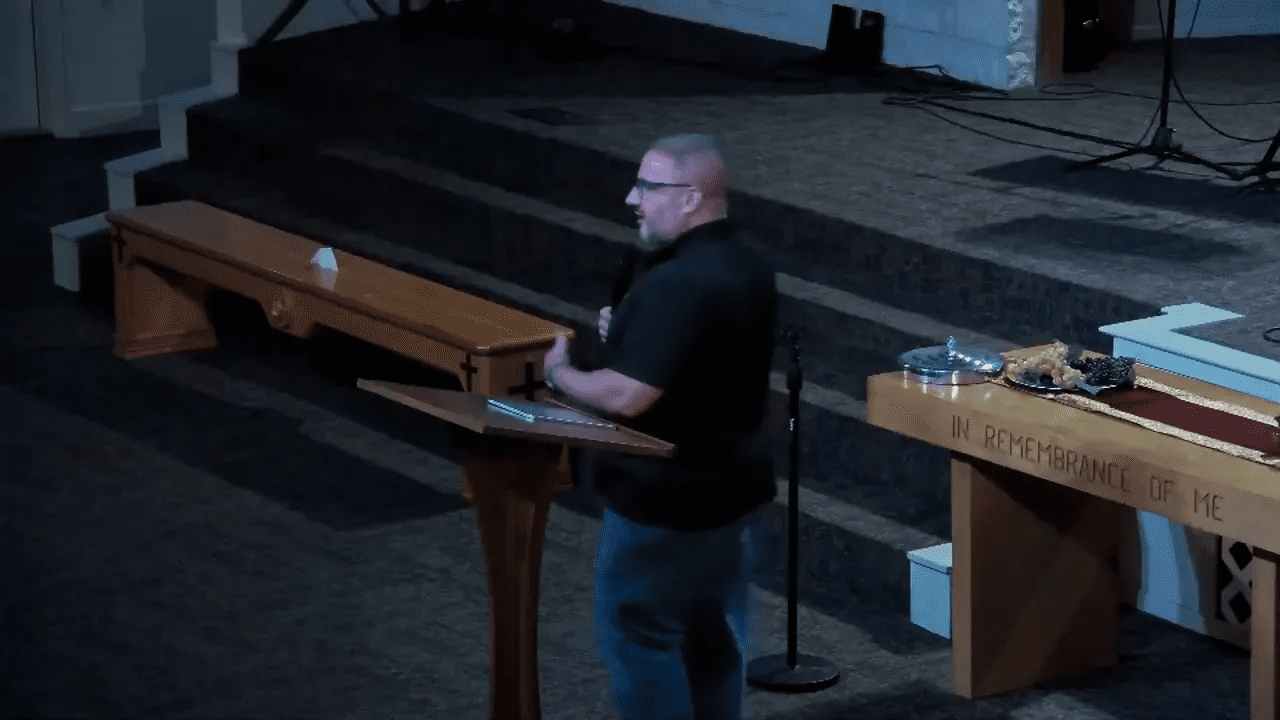Wide view of speaker at a lectern with a communion table and plates of grapes visible, altar inscription partially readable
