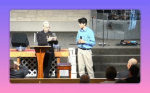 clear view of a speaker holding a mason jar on church stage with a young man beside him