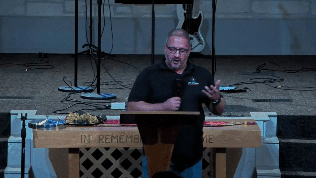 Frontal shot of a speaker at a lectern with communion elements on the table in front and stage equipment behind