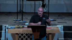 Frontal shot of a speaker at a lectern with communion elements on the table in front and stage equipment behind