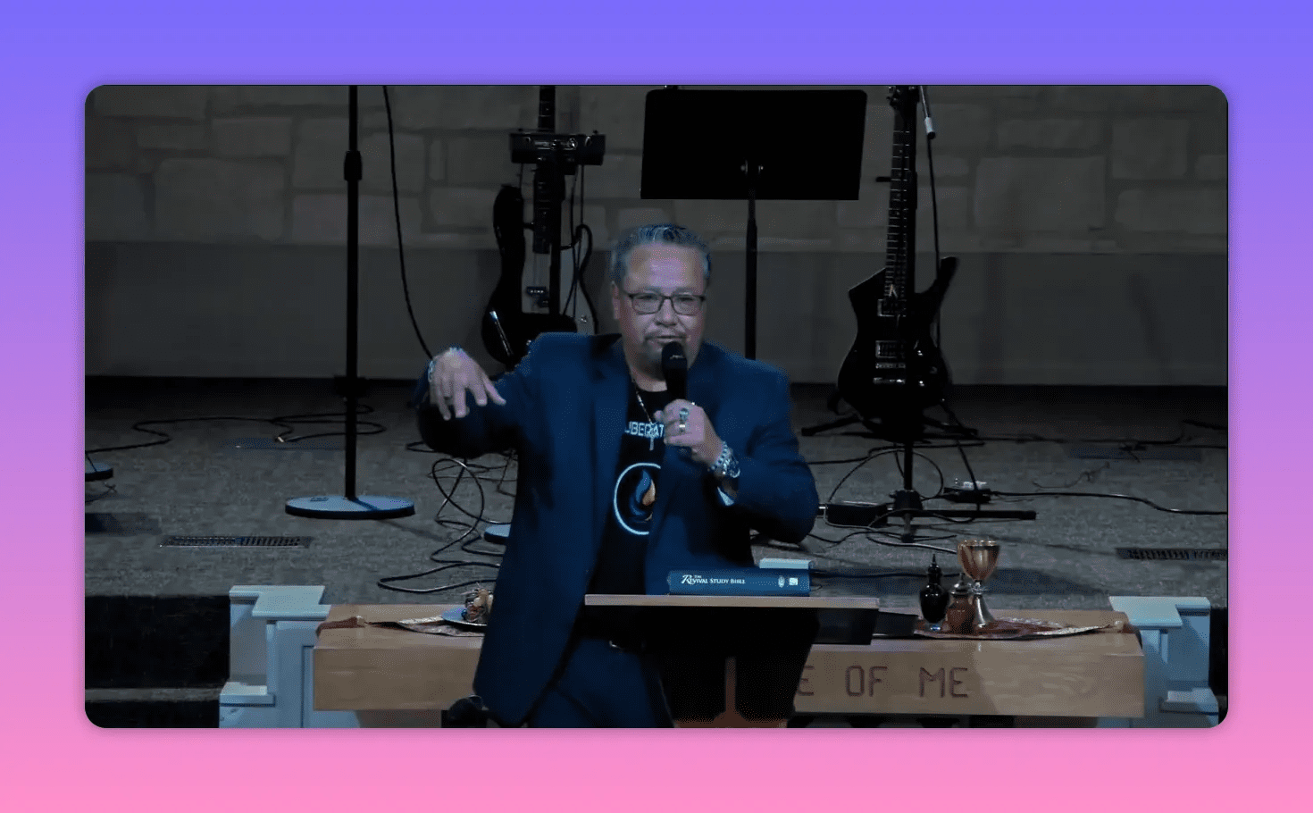 Centered view of the pastor at the pulpit speaking into a microphone with a hand gesture; Bible, communion cup and stage instruments visible behind him.