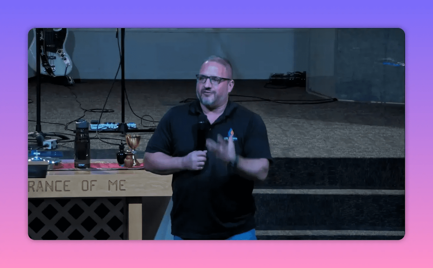 Church speaker on stage holding a microphone and using a hand gesture while addressing the congregation; communion table and steps visible