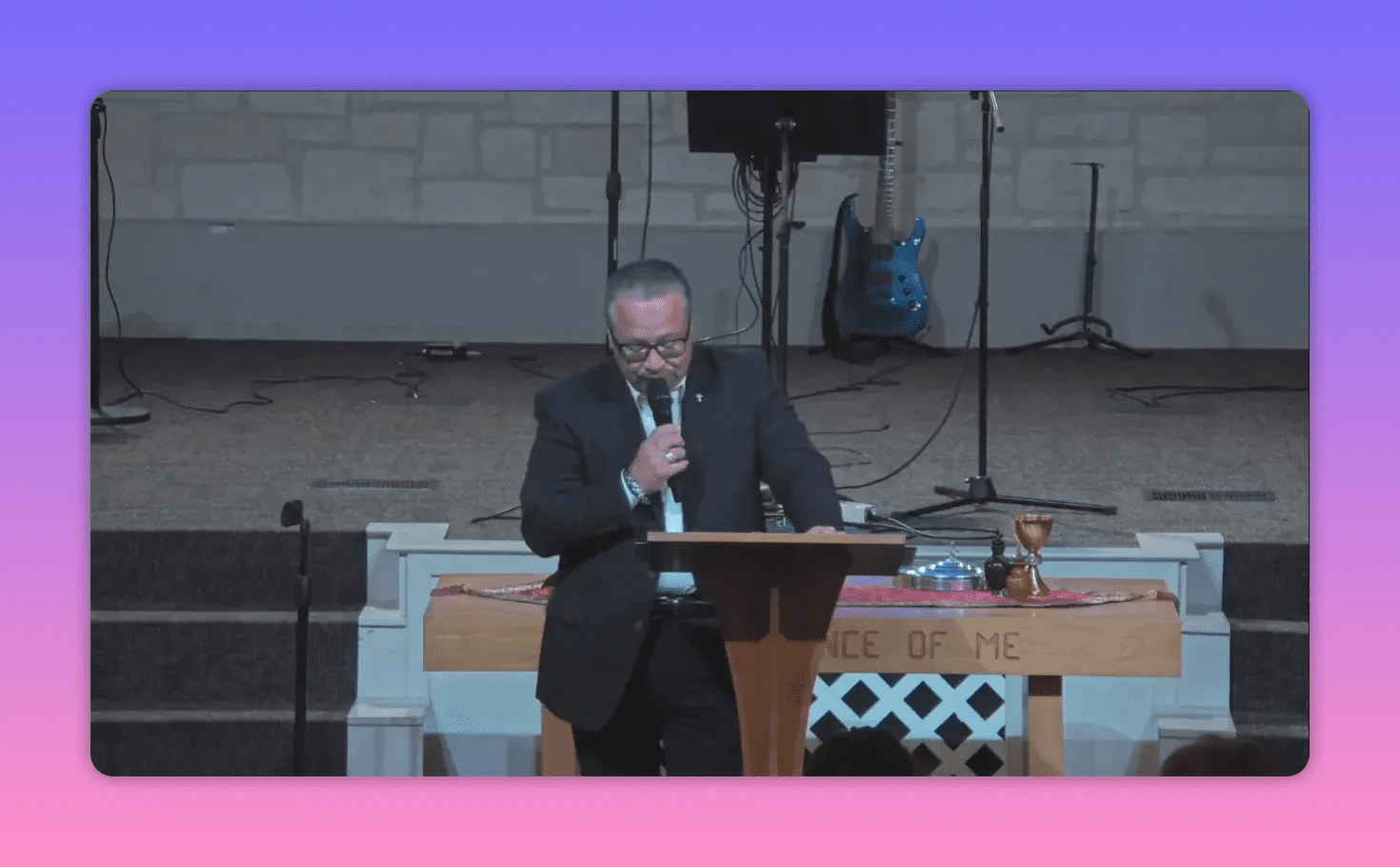 Speaker centered at a wooden lectern speaking into a microphone with communion elements on the table behind, well-lit and in focus.