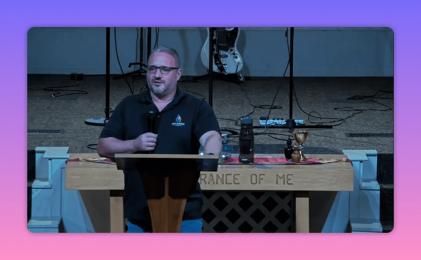 Church speaker centered behind a wooden lectern, microphone in hand, communion cup and water bottle visible on the table