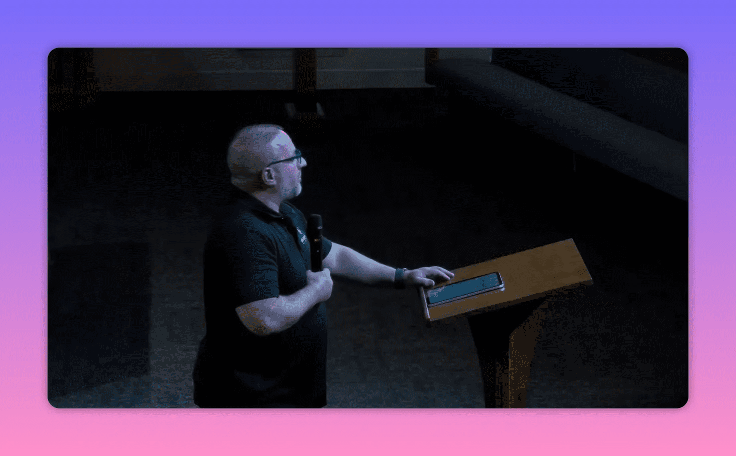 Speaker beside a lectern holding a microphone with one hand on a tablet, looking upward in a reflective pose