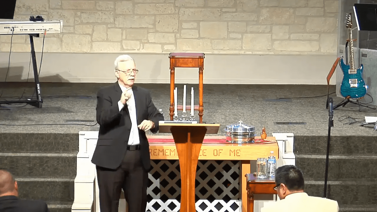 Pastor at pulpit gesturing while teaching, clear wide shot showing lectern, communion elements, and stage backdrop.
