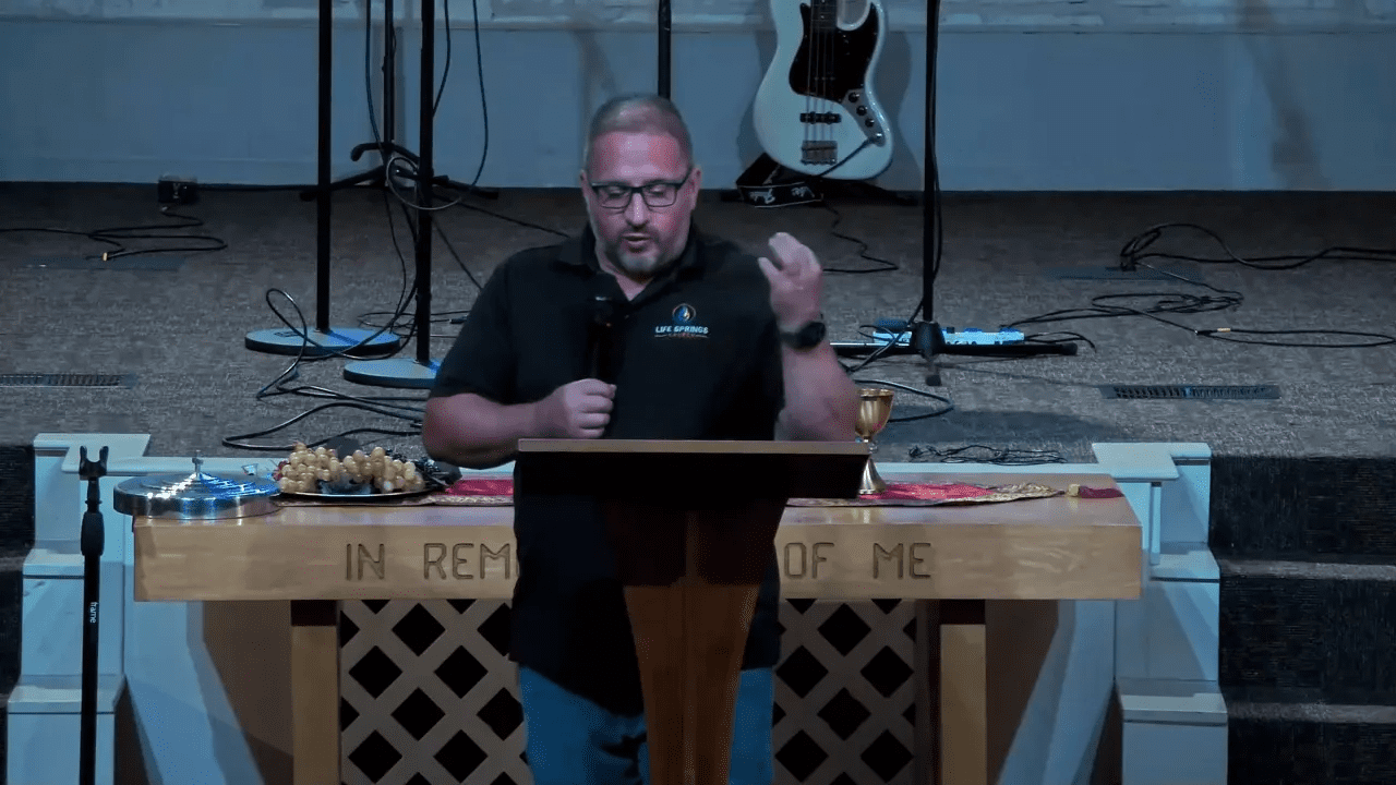 Frontal shot of a pastor speaking behind a communion table, holding a microphone with a chalice and grapes visible on the table