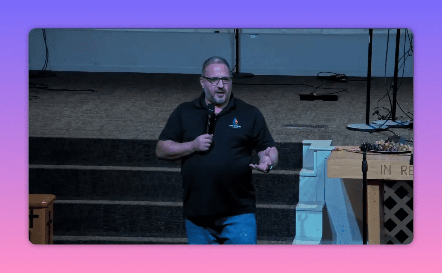 Speaker centered on a church stage holding a microphone and gesturing with one hand; communion table and stage equipment visible to the right