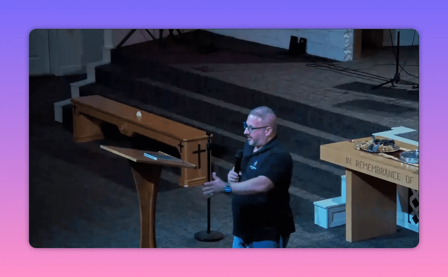 Church speaker standing beside a lectern, extending his arm while holding a microphone; communion table with elements and stage steps visible