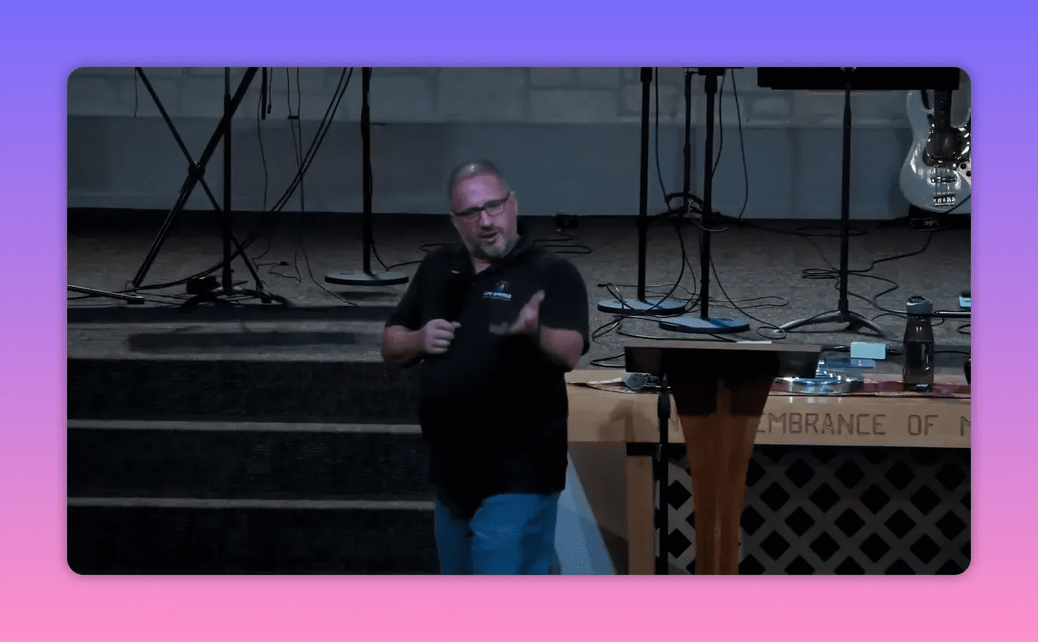 Speaker gesturing with an open hand while speaking beside the pulpit and communion table, stage instruments in background.