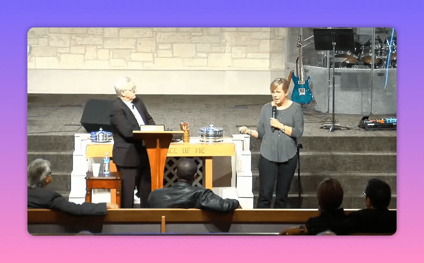 speaker holding a microphone and gesturing beside the pulpit with communion trays and stage instruments visible