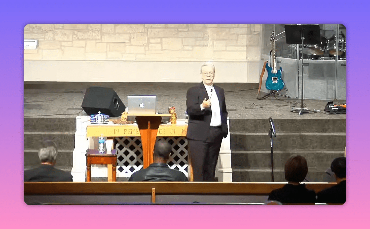 speaker standing to the right of pulpit with communion table, laptop and stage instruments visible