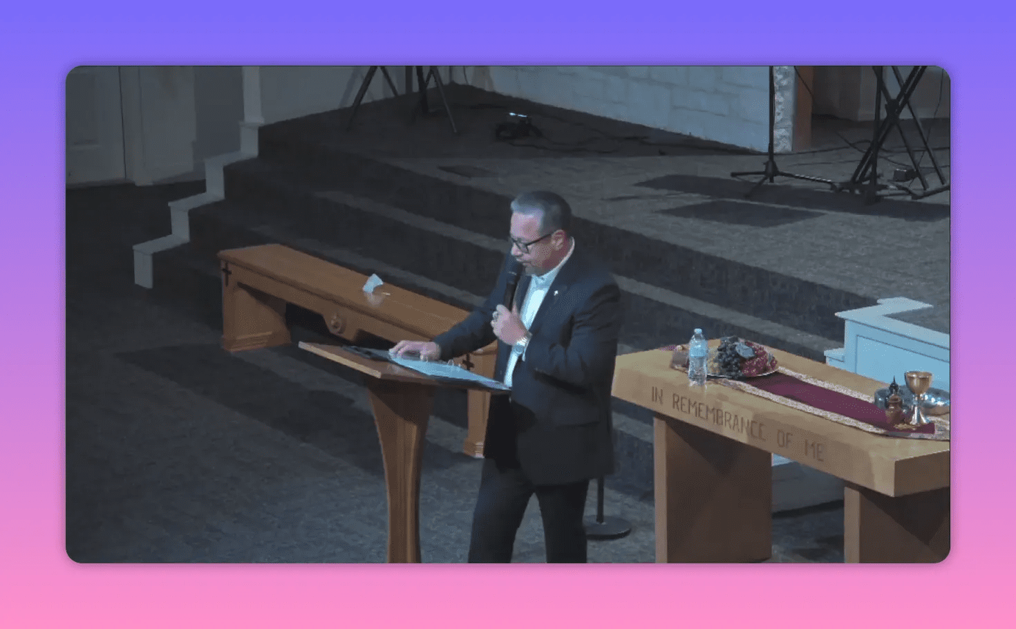 Pastor reading notes at a wooden lectern while holding a microphone, communion table with 'In Remembrance of Me' visible in front.