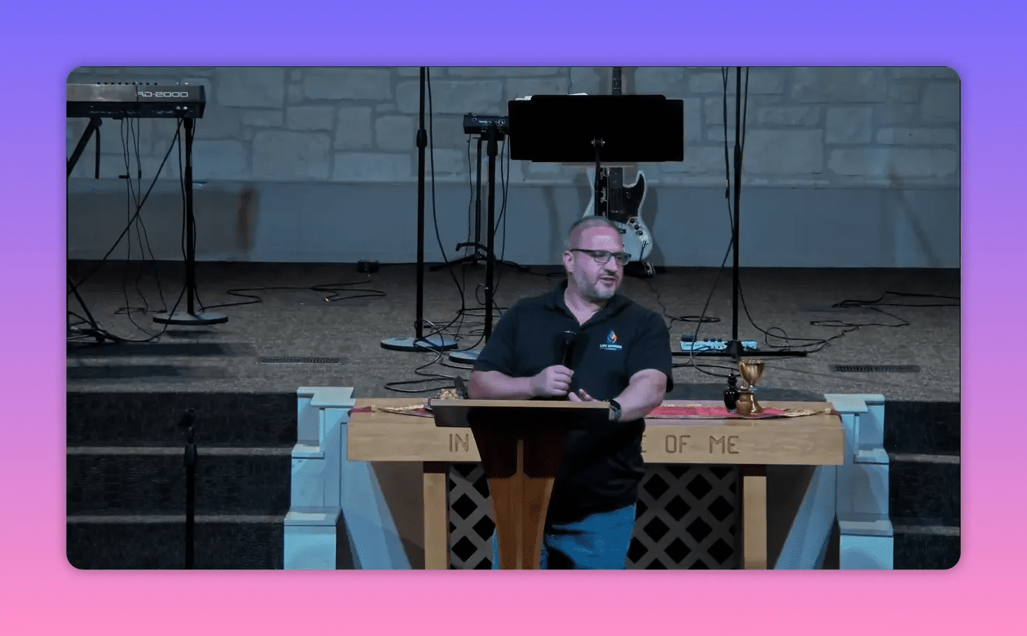 Church speaker at a wooden lectern holding a microphone and gesturing, altar inscription and stage instruments visible