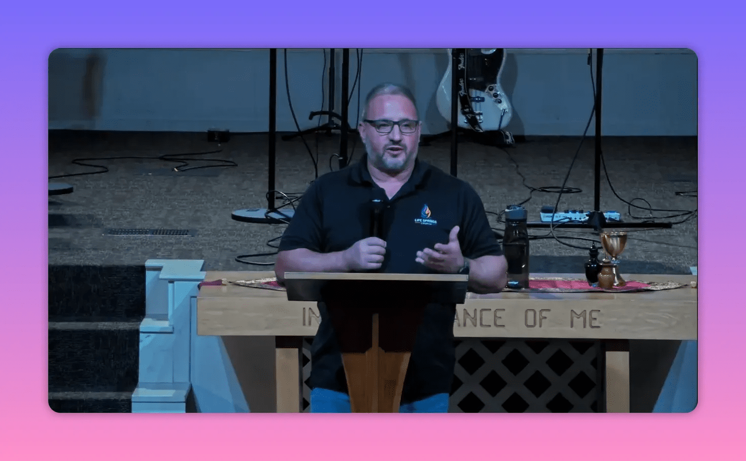 Church speaker centered behind a lectern, speaking with a hand gesture; guitar and stage gear visible behind him