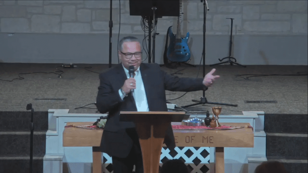 Speaker at a wooden lectern smiling and gesturing with an outstretched arm, communion elements visible on the table behind in a church sanctuary.
