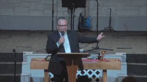 Speaker at a wooden lectern smiling and gesturing with an outstretched arm, communion elements visible on the table behind in a church sanctuary.
