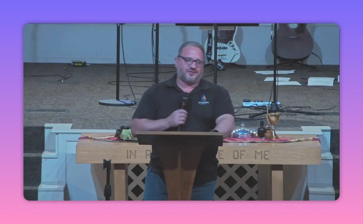 Speaker behind a lectern with communion elements on a table carved with 'In Remembrance Of Me' and guitars in the background