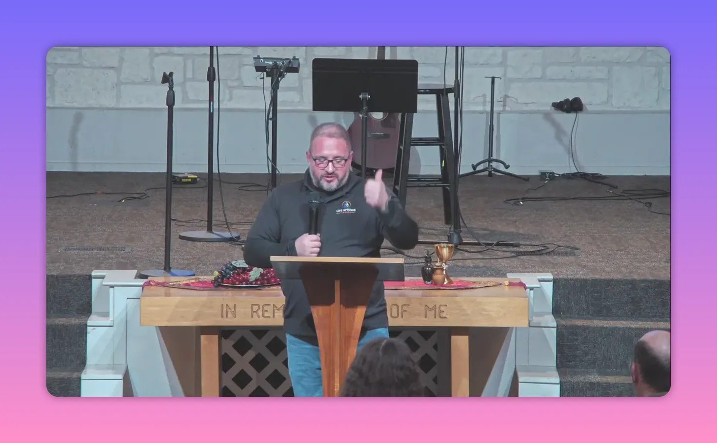 Pastor gesturing at a pulpit with communion elements on the table and the words 'IN REMEMBER OF ME' carved into the front.
