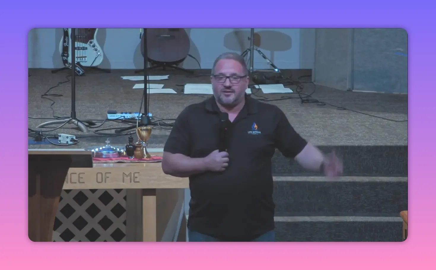 Speaker facing the congregation at the front of the church holding a microphone with communion elements and instruments on the table behind him