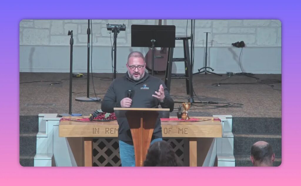 speaker gesturing at pulpit in church sanctuary with communion table and inscription visible