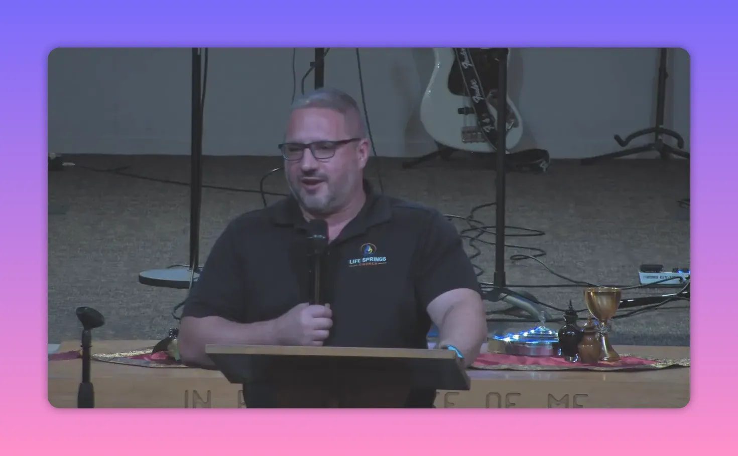 Speaker at a pulpit smiling while holding a microphone, communion chalice and plates visible to the right, stage instruments behind.