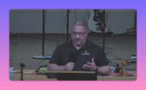 Speaker at a pulpit holding a microphone and gesturing with one hand; communion cups and plates visible on the table behind, guitar in background.