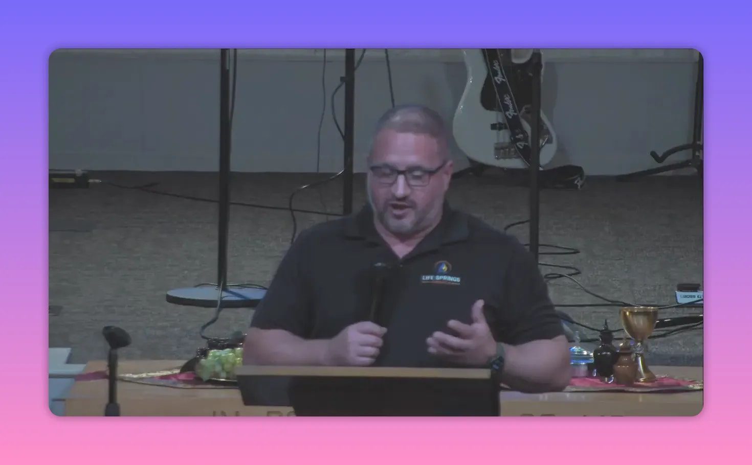 Speaker at a pulpit holding a microphone and gesturing with one hand; communion cups and plates visible on the table behind, guitar in background.
