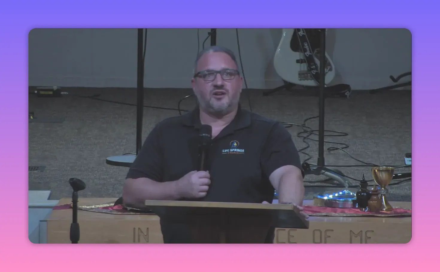 Speaker at a pulpit facing forward and speaking into a microphone, communion cups and plates visible on the table beside him and musical instruments behind.