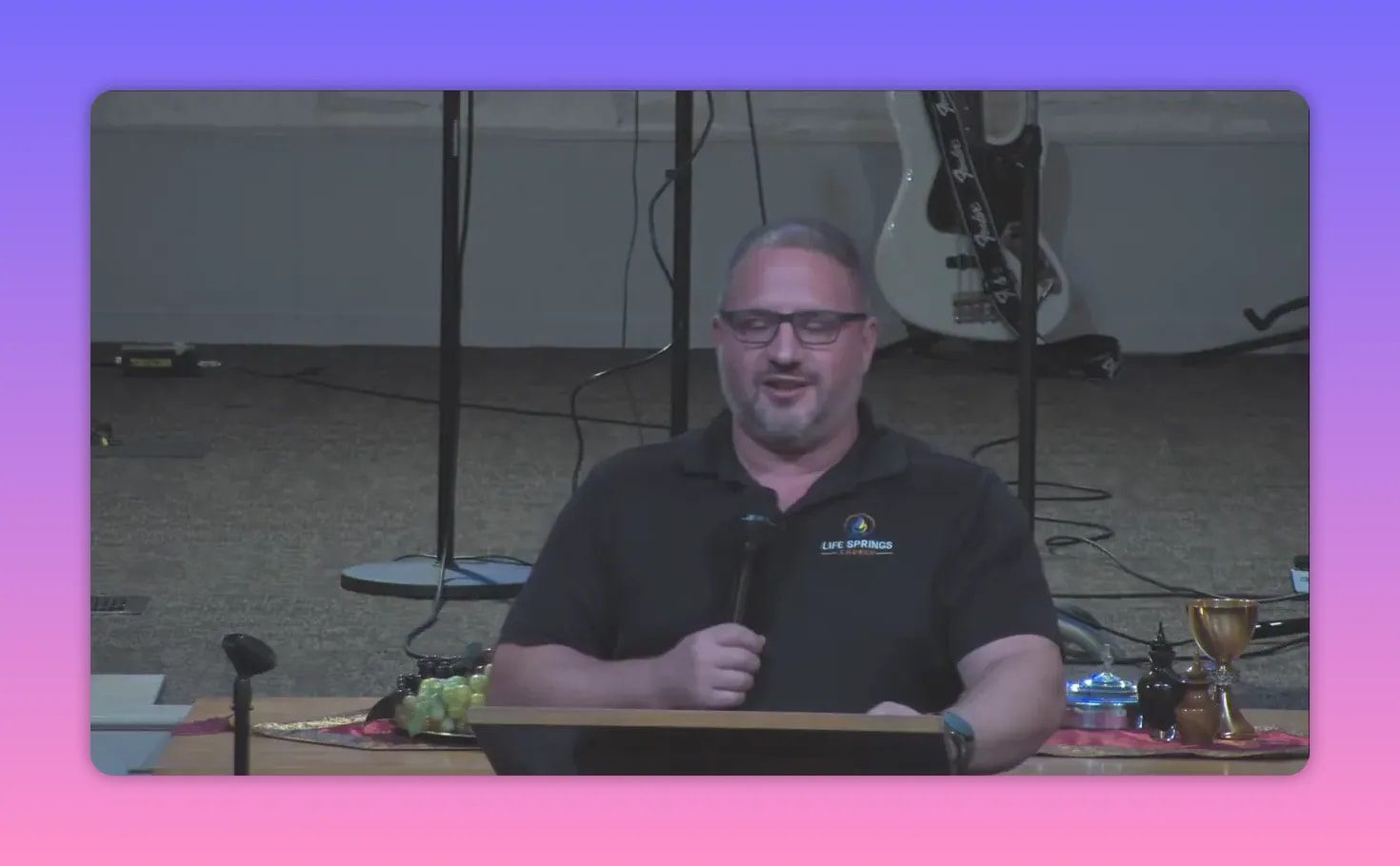 Centered view of a speaker at a pulpit holding a microphone, communion grapes and cups on the table beside him, guitar and mic stands behind.