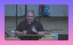 Church speaker at a lectern making a clenched-hand gesture while speaking, with a guitar in the background