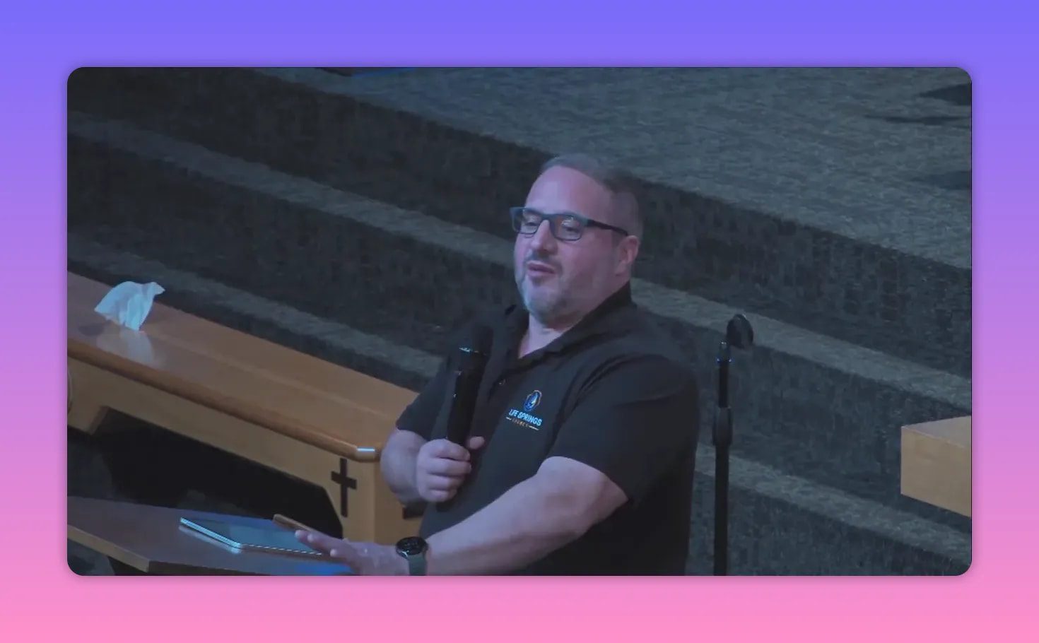 Frontal view of a speaker at a lectern gesturing with one hand while holding a microphone, church steps behind