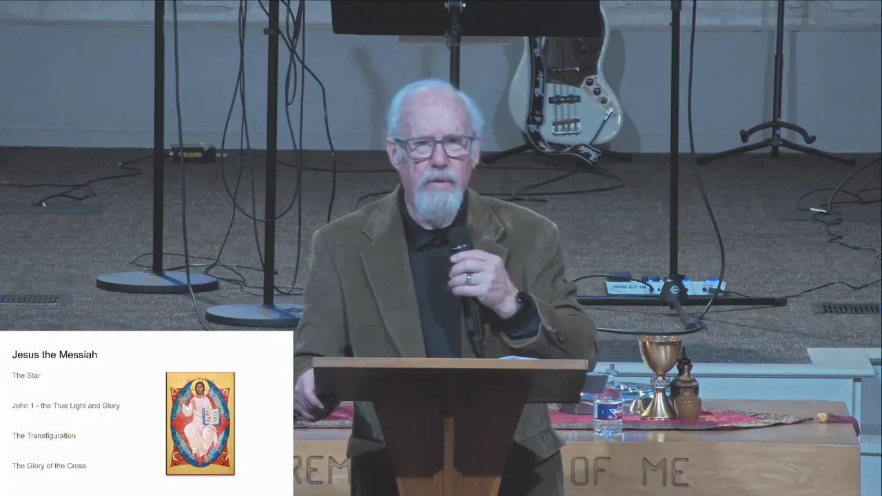Clear frontal image of the speaker at a podium holding a microphone with a presentation slide titled 'Jesus the Messiah' visible in the lower-left and communion vessels on the table.