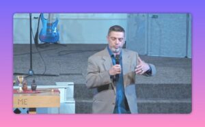 Speaker on stage holding a microphone and gesturing with his hand, clear view of steps and guitar in background