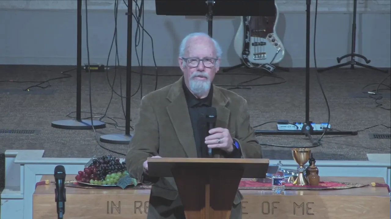 Speaker facing the camera at a pulpit holding a microphone, communion elements visible on the table behind