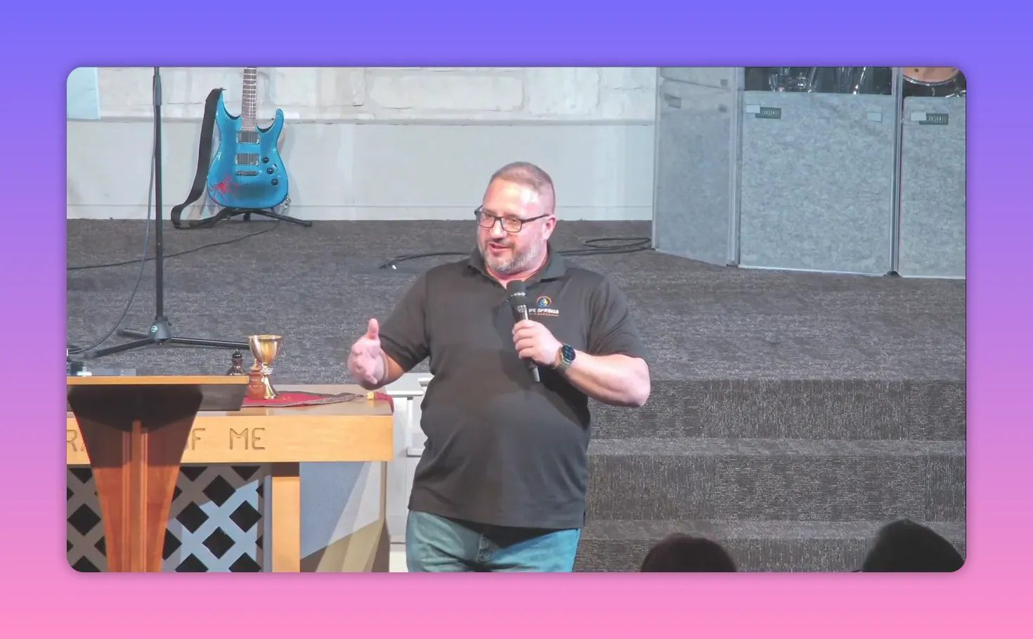 Speaker on stage beside the communion table with a chalice; the carved words 'IN REMEMBRANCE' are visible on the table front.