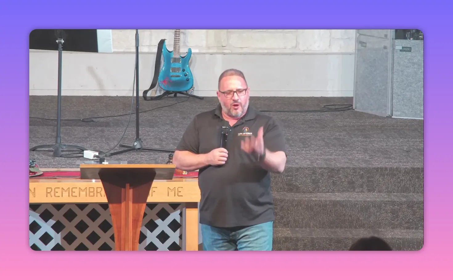 pastor standing by the pulpit with hand extended, communion table front reading 'IN REMEMBRANCE OF ME' clearly visible
