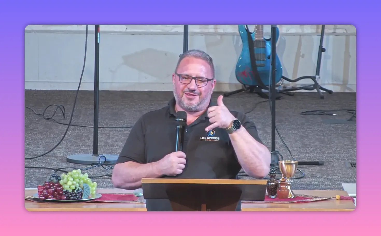 Outdoor church speaker holding a microphone and pointing toward the audience at an outdoor stage lectern.